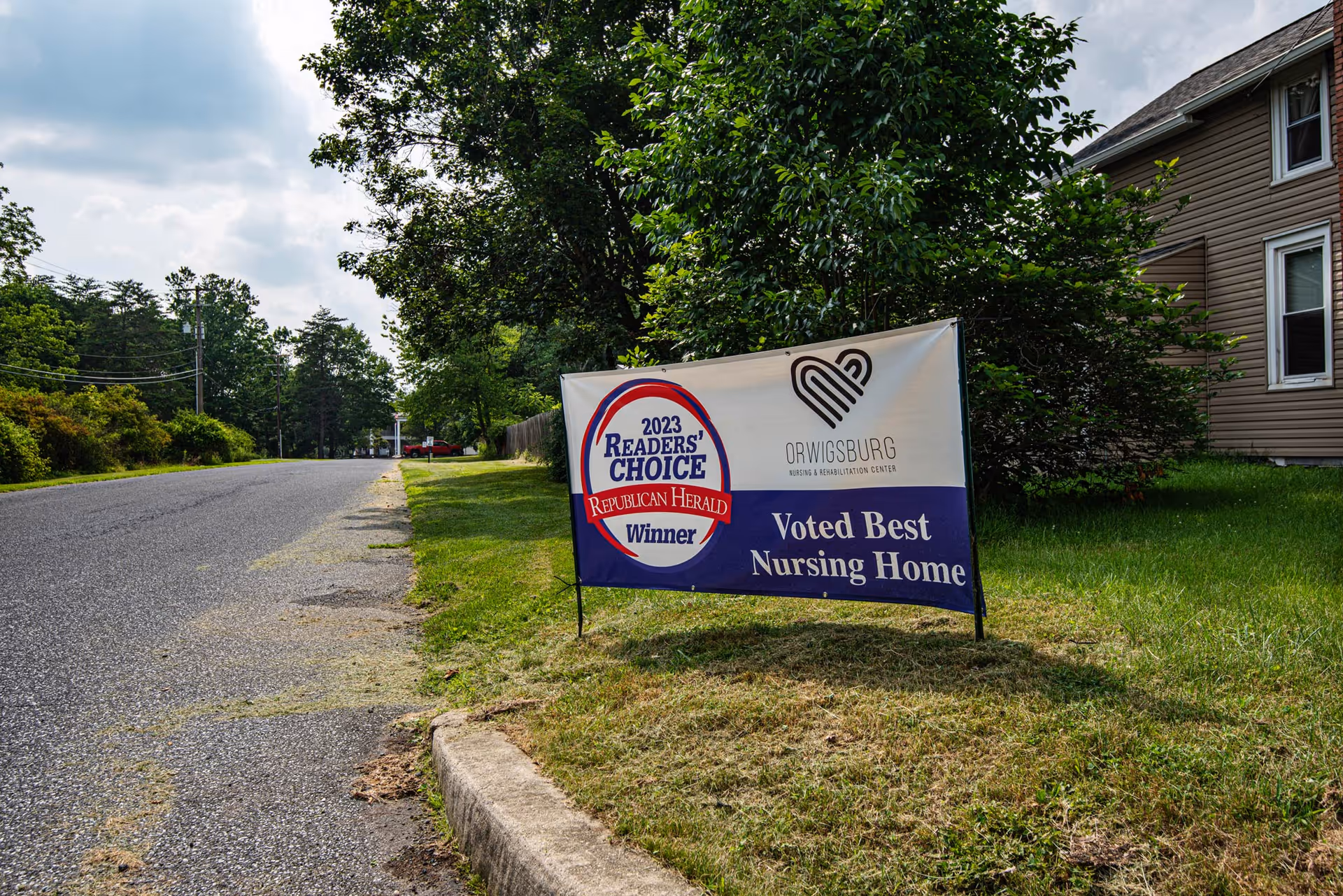 A roadside lawn with a sign that reads '2023 Readers' Choice Republican Herald Winner, Orwigsburg Nursing & Rehabilitation Center, Voted Best Nursing Home'. Trees and a house are visible in the background under a partly cloudy sky.
