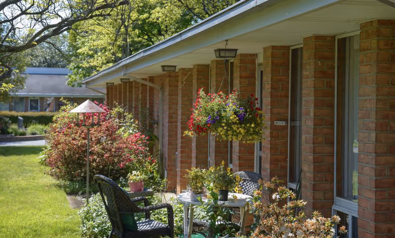 Outdoor patio area at Friends House Retirement Community featuring a brick building with hanging flower baskets, a small round table with potted plants, and two black wicker chairs surrounded by greenery and bushes under a large tree.