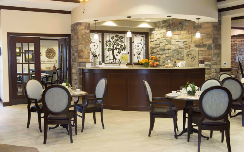 A cozy dining area in a senior living facility with round tables and cushioned chairs. The room features a stone accent wall behind a dark wood counter with pendant lights hanging above. Decorative stained glass with a tree design is visible behind the counter. The floor is light-colored, and there are flowers and table settings on the tables. A doorway with glass-paneled double doors leads to another room in the background.