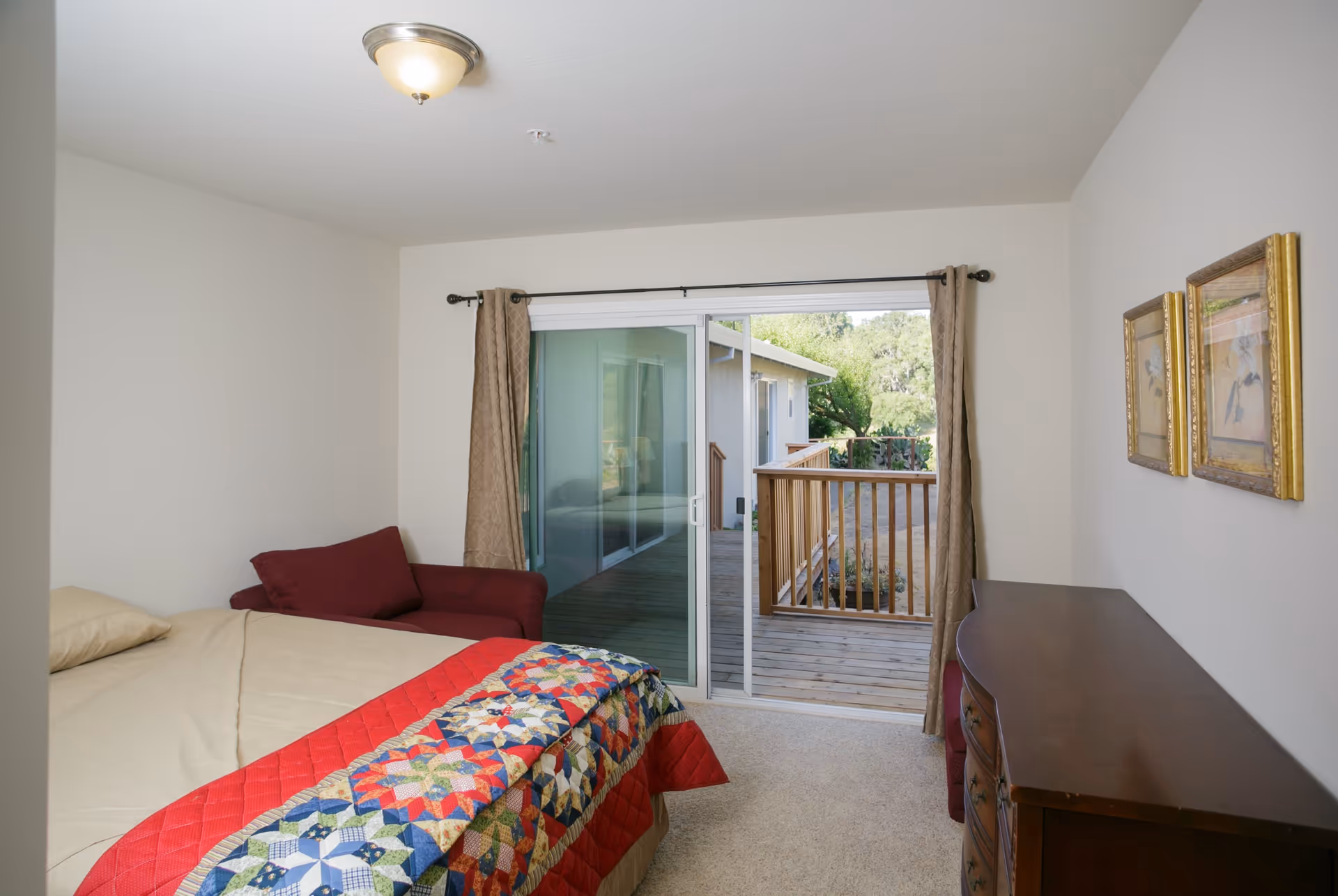 Bedroom with a bed covered by a colorful quilt, a red armchair, a wooden dresser, and a sliding glass door opening onto a small wooden deck.