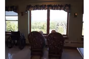 Two patterned armchairs facing a large window with a scenic view of trees and hills outside. A wheelchair is positioned near the window on the left side. The room has light-colored walls and a floral valance above the window.