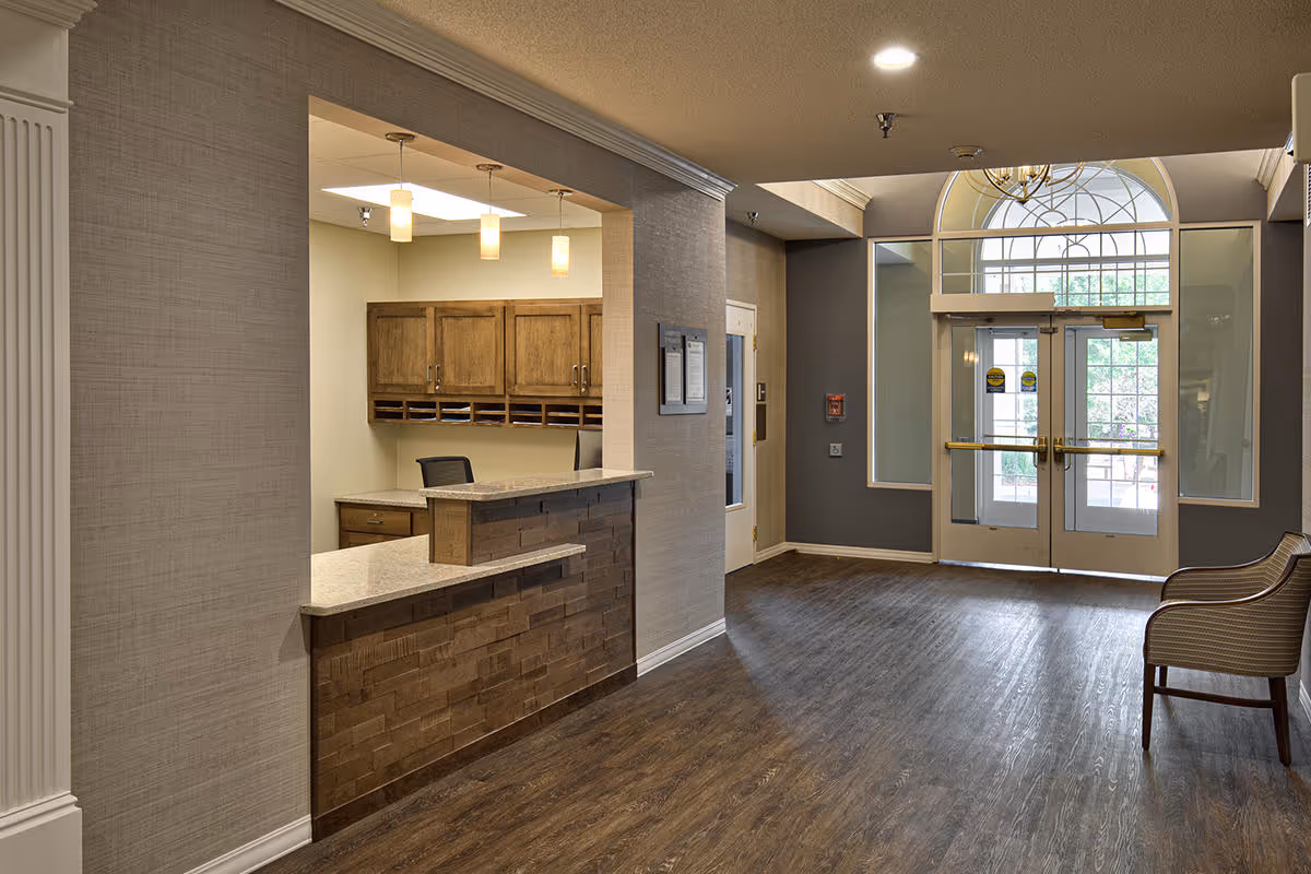 Interior view of a senior living facility lobby area with a reception desk on the left featuring a granite countertop and wooden cabinetry behind it. The floor is wood, and there are double glass doors with windows above them letting in natural light. Two chairs are placed on the right side near the entrance.
