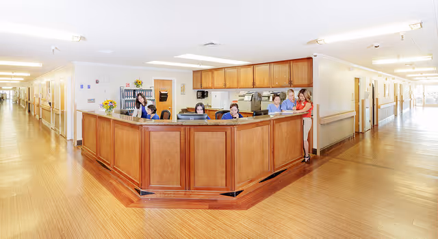 Reception area in a senior living facility with a wooden front desk and several staff members behind it. The area is well-lit with overhead lights, and there are long hallways extending on both sides of the desk. The floor is wooden, and the walls are light-colored with handrails along the hallways.