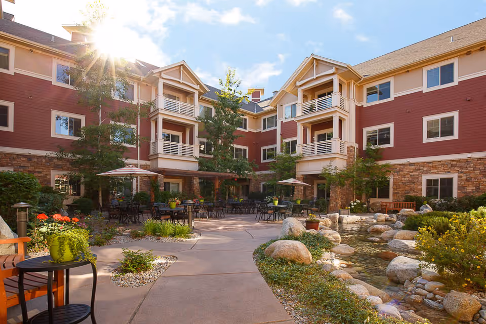 Sunlit courtyard with tables, umbrellas, landscaping and a small pond in front of a three-story red and stone-clad residential building.