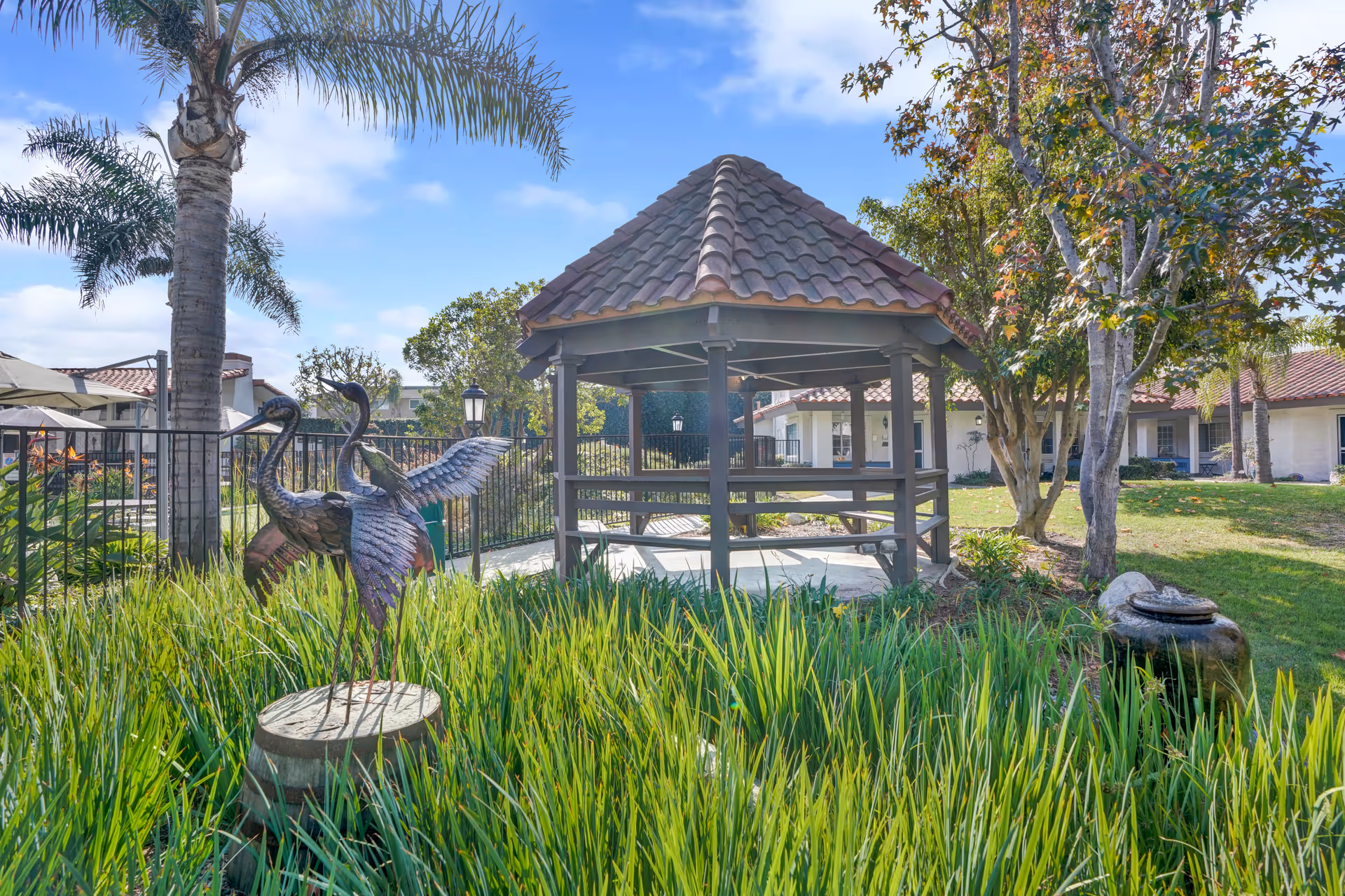 Outdoor garden area at Huntington Terrace featuring a gazebo with a tiled roof, surrounded by green grass and trees. In the foreground, there is a metal sculpture of two cranes standing on a wooden stump. The sky is clear with a few clouds.