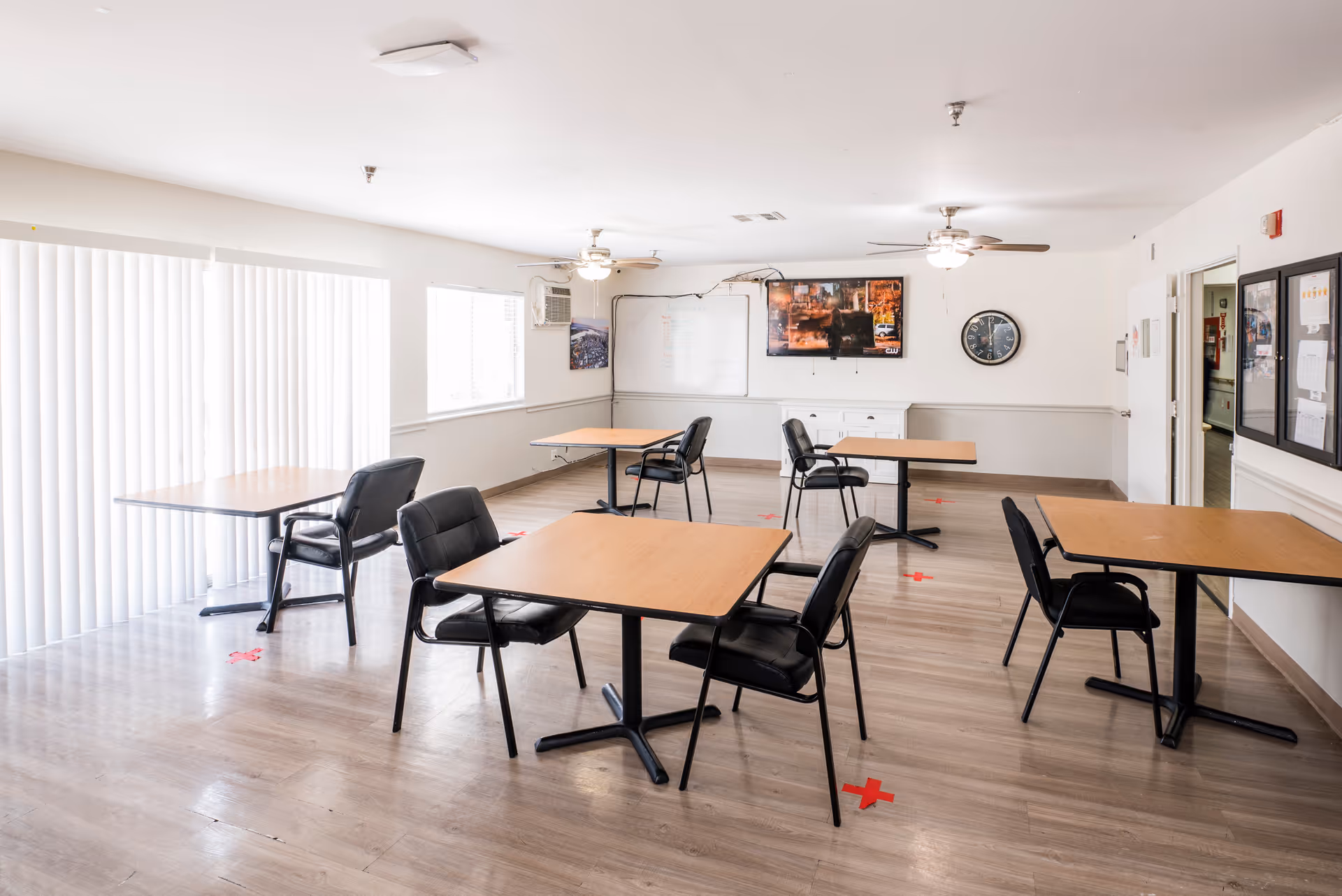 A bright and clean dining room with several wooden tables and black chairs spaced apart. The room has light-colored walls, large windows with vertical blinds, ceiling fans with lights, a wall clock, a whiteboard, and a mounted TV. Red tape marks on the floor indicate social distancing measures.