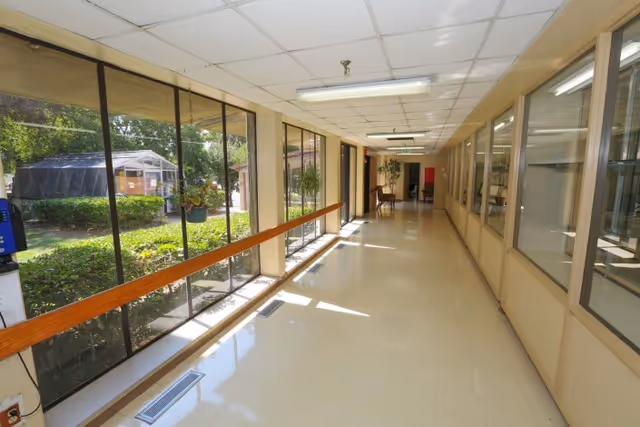 A bright hallway with large windows on one side overlooking a garden area with greenery and a greenhouse. The opposite side has glass windows looking into an interior room. The floor is shiny and light-colored, and there are handrails along the windowed wall. Fluorescent lights are on the ceiling.