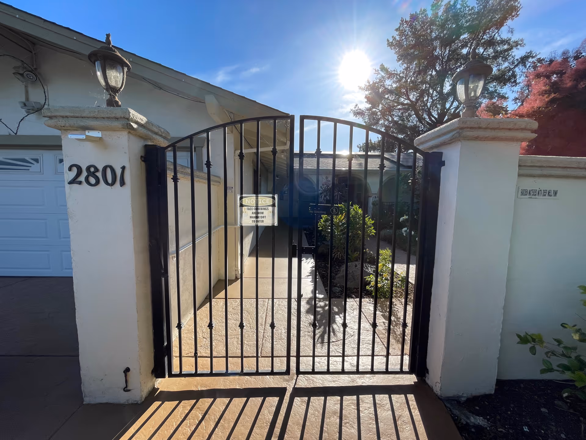 Black metal gate entrance with the number 2801 on a white pillar to the left and a white wall on the right. There are two lantern-style lights on top of the pillars. Behind the gate, there is a pathway leading to a building entrance with plants on either side. The sun is shining brightly in the blue sky.