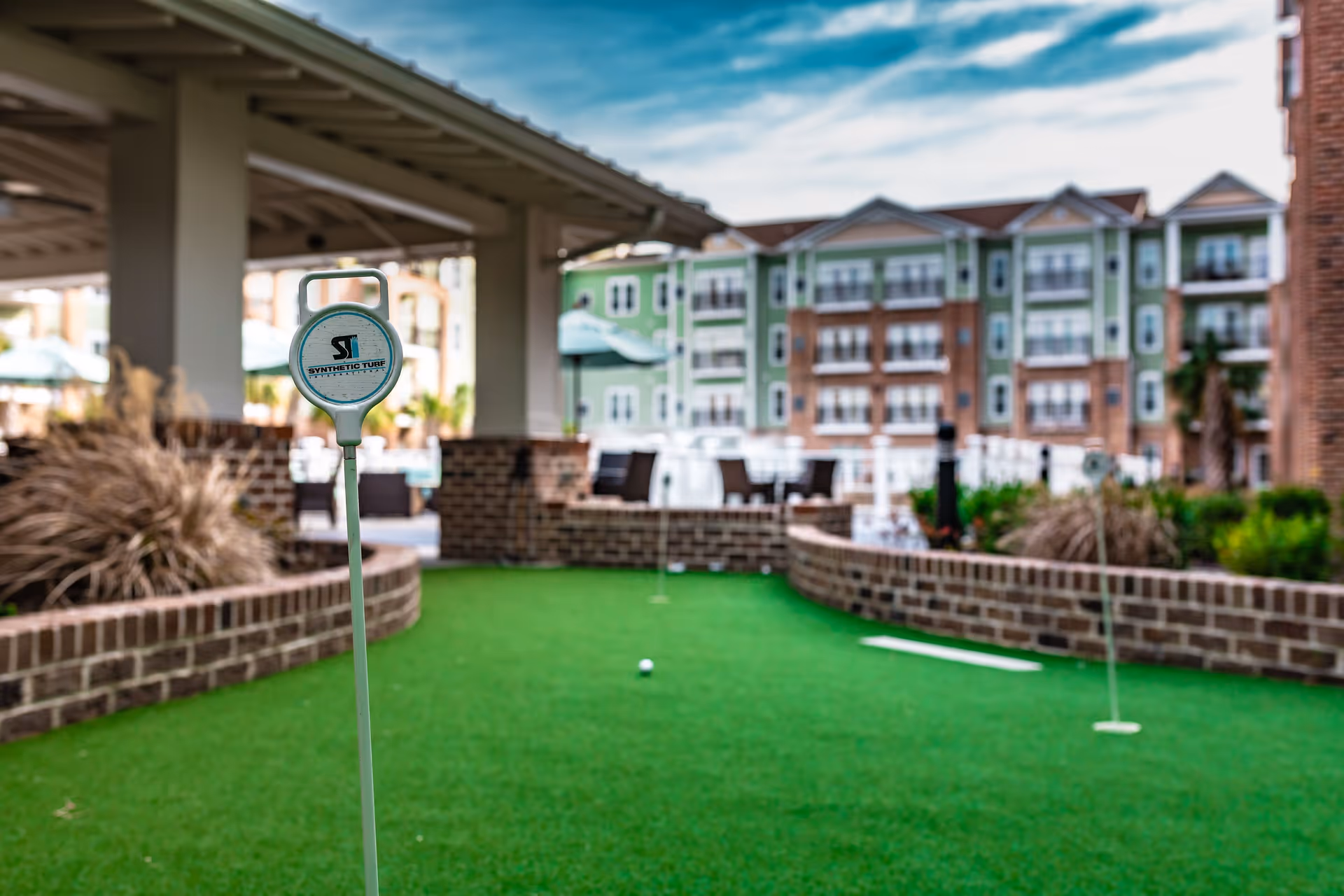 Outdoor putting green with synthetic turf at a senior living facility, surrounded by brick walls and landscaping, with multi-story residential buildings in the background under a partly cloudy sky.