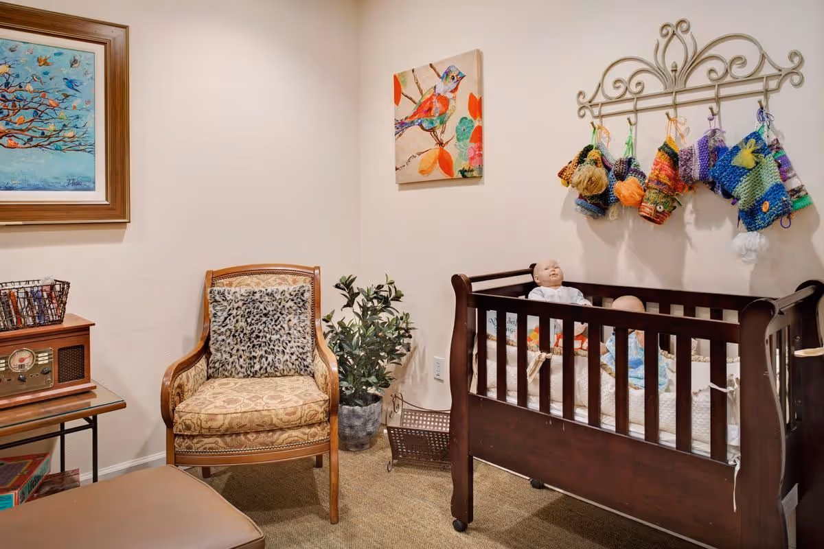 A cozy room with a wooden crib containing two baby dolls, a vintage armchair with a leopard print cushion, a small table with a retro radio, a potted plant, and colorful artwork on the walls.