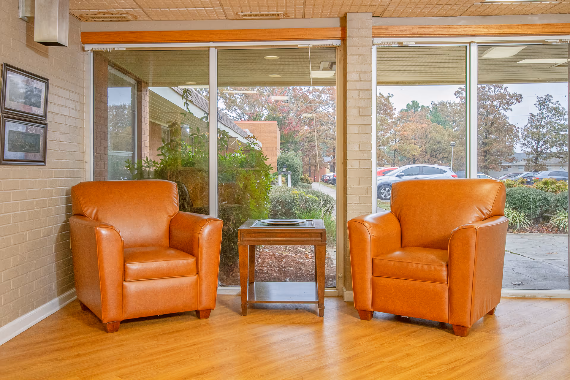 Two orange leather armchairs with a small wooden table between them, placed in front of large floor-to-ceiling windows showing an outdoor view with trees and parked cars.