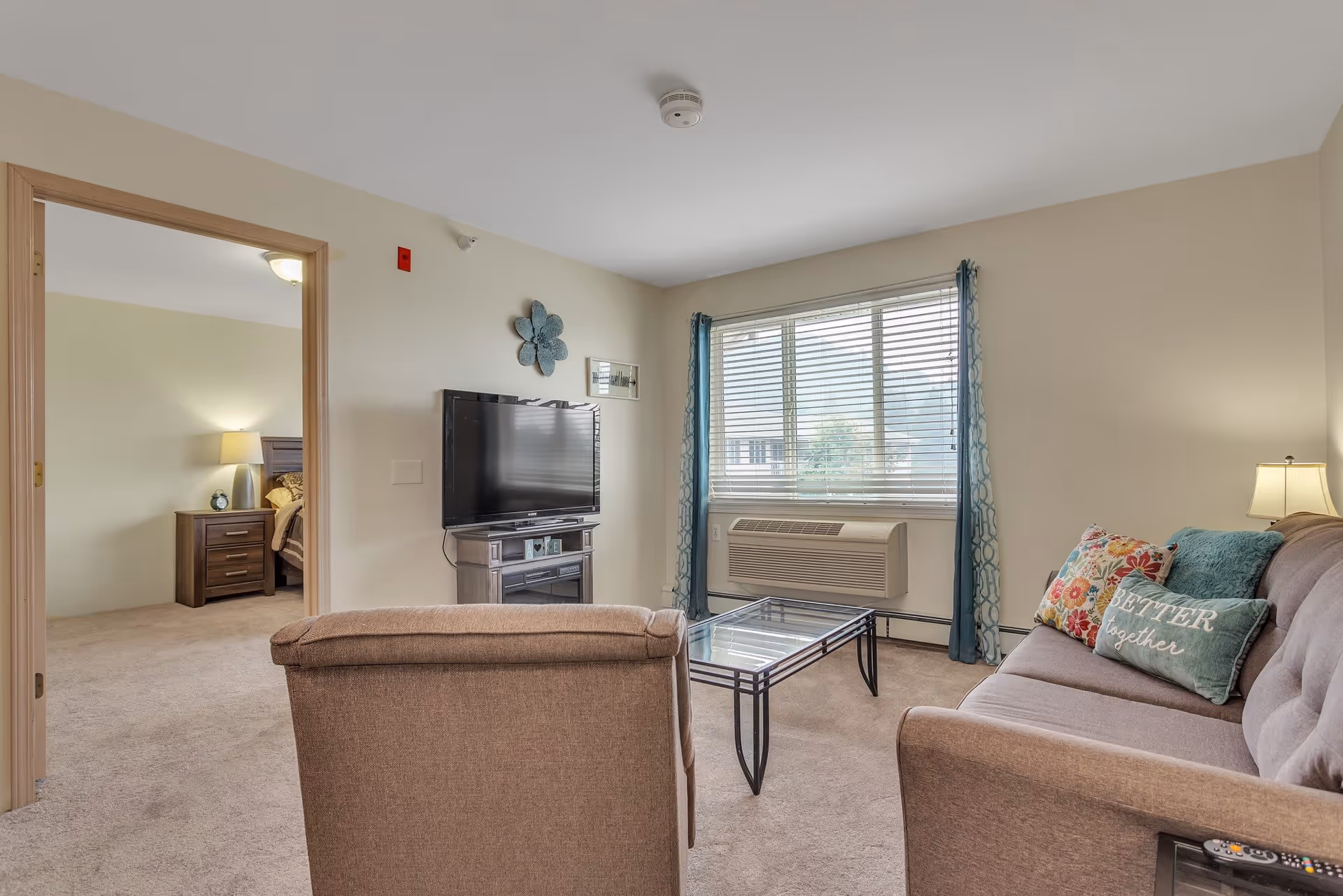 A cozy living room with a beige armchair and a sofa adorned with colorful pillows, including one that says 'Better together'. A glass coffee table sits in front of the sofa. A flat-screen TV is mounted on a stand against the wall. The room has a large window with blue patterned curtains and an air conditioning unit below it. Through an open doorway, a bedroom with a nightstand and lamp is visible.