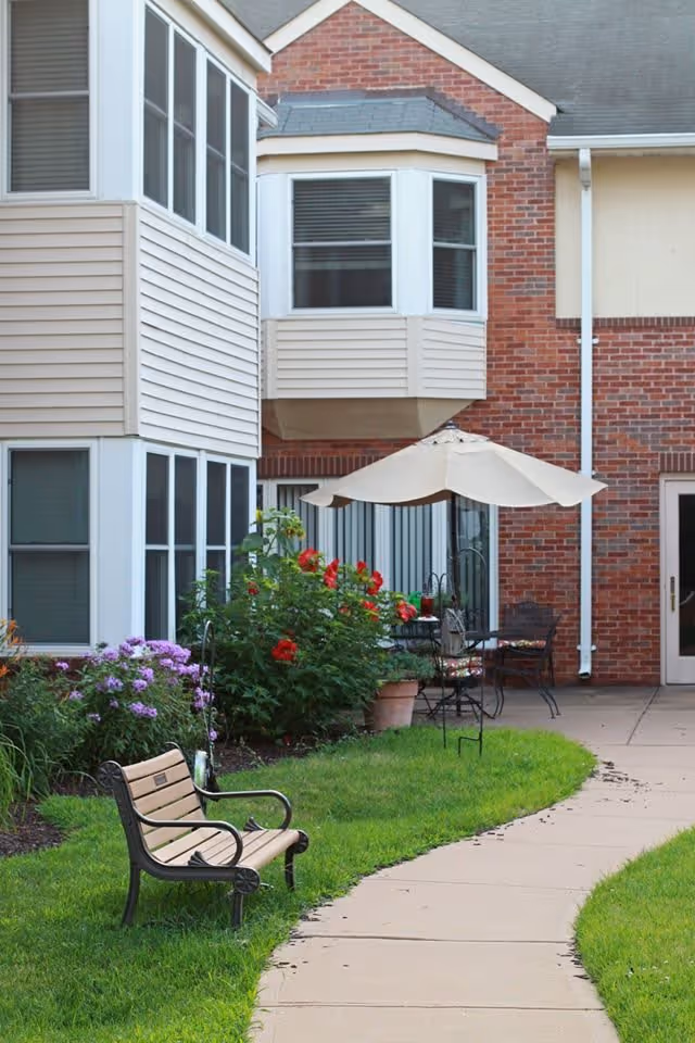Outdoor patio area at Brooking Park featuring a curved concrete walkway, a wooden bench with metal armrests, green grass, flower bushes with red and purple flowers, and a table with chairs under a large beige umbrella next to a brick and siding building.