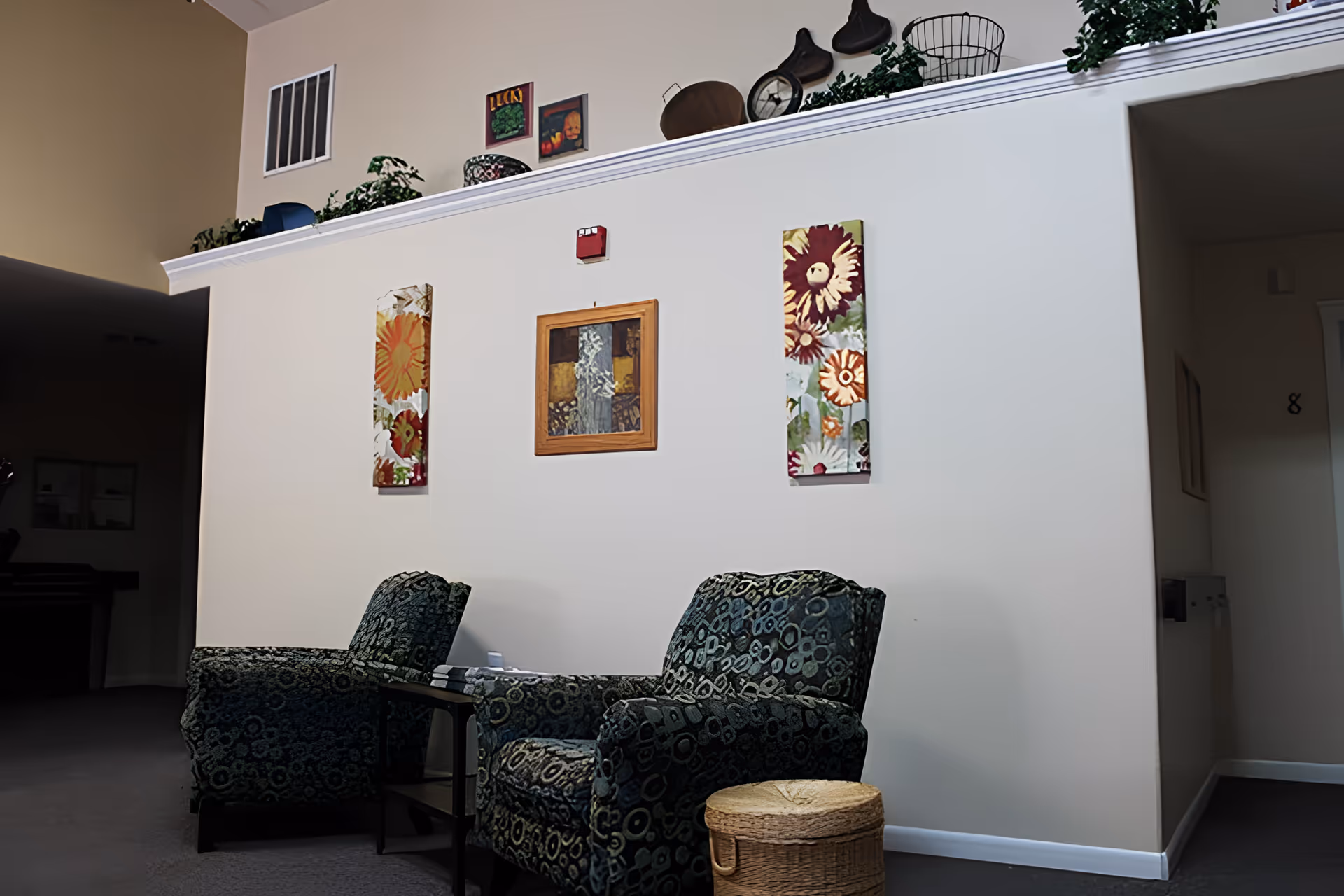 A cozy seating area with two patterned armchairs and a small side table between them. The wall behind the chairs is decorated with three pieces of artwork featuring floral and abstract designs. Above the wall is a ledge with various decorative items including baskets, a clock, and greenery. The room has a neutral color palette with beige walls and carpeted floor.