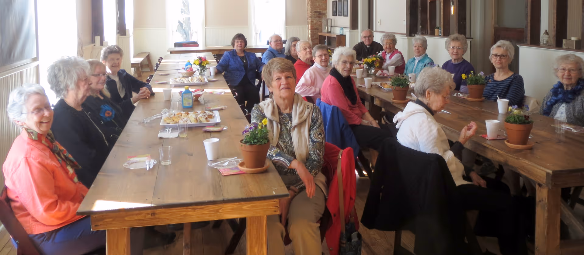A large group of elderly residents seated around long wooden tables in a bright communal dining/activity room with potted plants and refreshments.