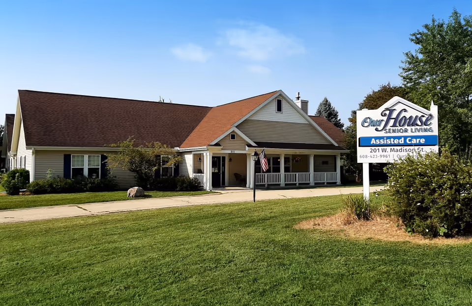 Exterior view of Our House Senior Living - Cambridge Assisted Care building with a large front porch, an American flag, and a sign displaying the facility's name and address. The building has beige siding, a brown roof, and is surrounded by green grass and some bushes under a clear blue sky.