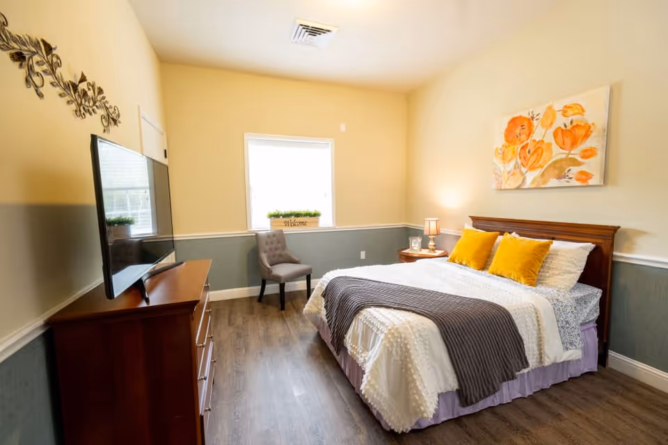 Well-lit, neatly furnished bedroom with a bed, bedside table and lamp, TV on a dresser, chair by the window, and floral wall art.