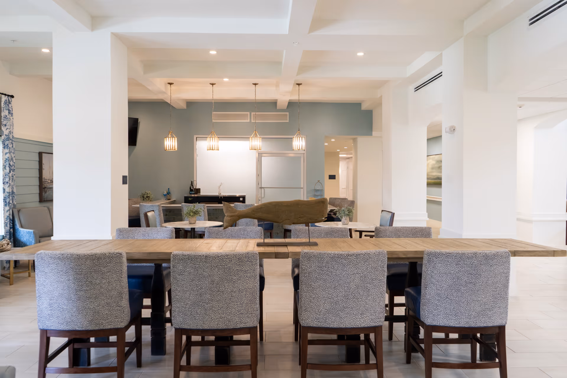 Bright communal dining and lounge area with a long wooden table surrounded by upholstered chairs, pendant lights, and seating in the background.