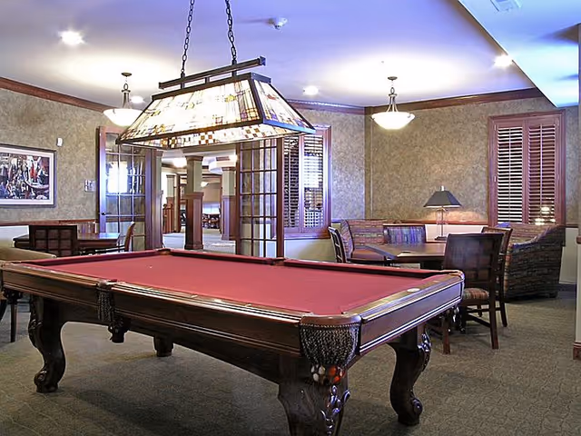 Interior room with a red felt pool table in the foreground, a stained glass hanging light fixture above it, and seating areas with chairs and a couch in the background. The room has beige walls, wooden window shutters, and carpeted floors.