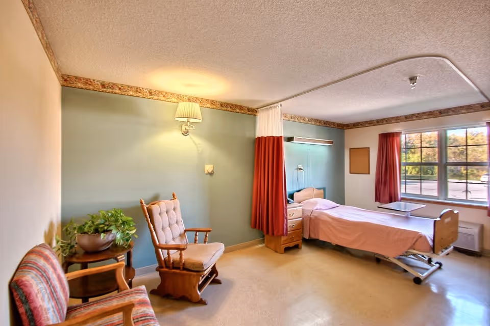 A bright senior-care bedroom with a single adjustable bed, bedside furniture, a privacy curtain, chairs, and a window.