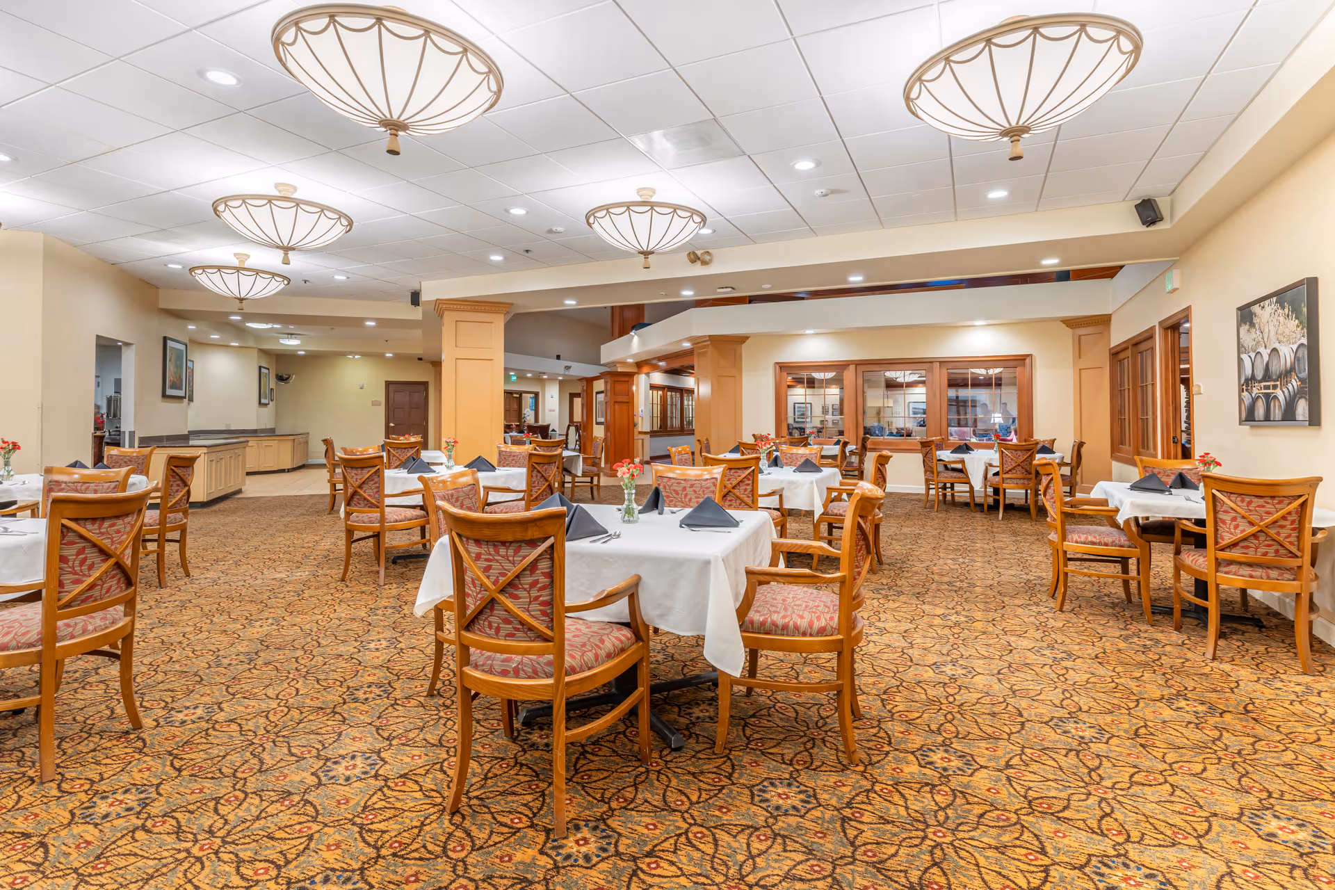 Spacious dining room with tables set with white tablecloths, wooden chairs, patterned carpet, and decorative ceiling lights.