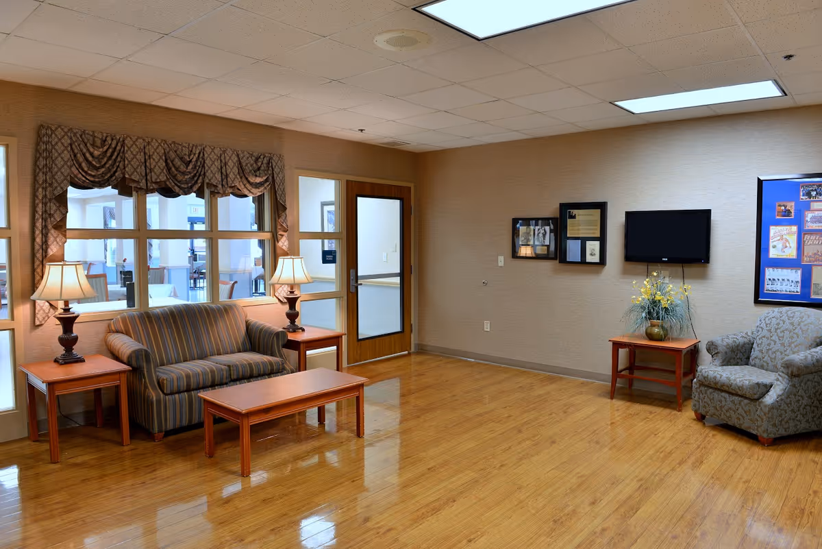Furnished common living area with a striped loveseat, armchair, coffee and side tables, lamps, wall-mounted TV, and bulletin board on a polished wood floor.