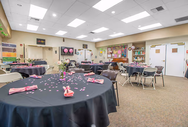 Community dining/activity room with round tables draped in black tablecloths, pink napkins and confetti, folding chairs, and decorated bulletin boards on the walls.
