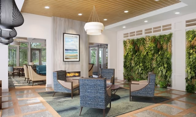 A modern senior living community common area featuring a cozy seating arrangement with four blue upholstered armchairs around a circular coffee table on a green rug. The room has a contemporary fireplace with a framed landscape painting above it. On the right wall, there is a vertical garden with lush green plants. Large windows and glass doors allow natural light to fill the space.