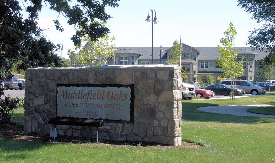 Stone entrance sign for Middlefield Oaks Senior Living Community on a lawn with the facility building and parked cars visible behind it.