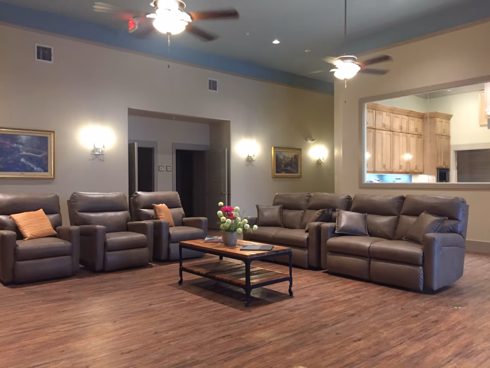 A spacious living room with three gray recliner chairs and two gray sofas arranged around a wooden coffee table with a flower vase on it. The room has wooden flooring, beige walls with framed paintings, wall sconces, and two ceiling fans with lights. A large window opening shows a kitchen area with wooden cabinets.