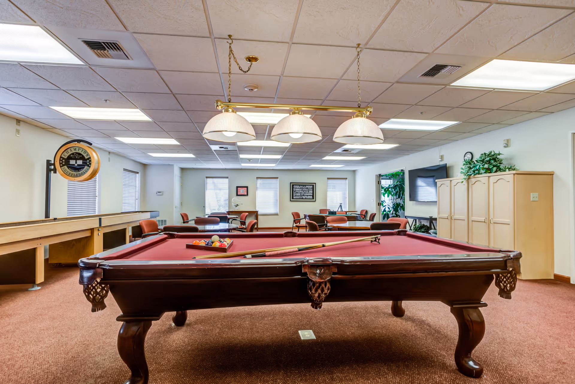 A recreational room with a red felt pool table in the foreground, pool cues and balls arranged on it. Behind the pool table, there are several tables and chairs arranged for seating. The room has a carpeted floor, fluorescent ceiling lights, and a shuffleboard table along the left wall. There is a large TV mounted on the right wall above a set of cabinets, and some plants are placed near the TV. Windows with blinds allow natural light into the room.
