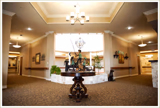 Spacious, well-lit interior lobby with a round wooden table topped by a sculpture and plants, chandeliers overhead, and carpeted hallways leading off to each side.