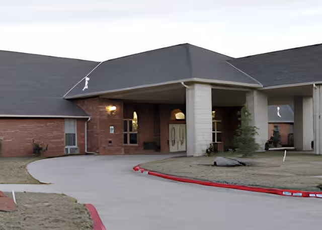 Exterior view of the entrance to a single-story brick building with a covered driveway and a sloped roof, surrounded by a paved driveway and some grass areas.