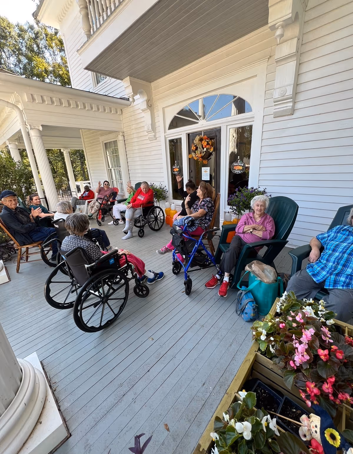 A group of elderly people sitting and socializing on the porch of a white building with columns. Some are in wheelchairs, others in chairs, surrounded by potted plants and flowers. The porch has a wooden floor and a decorative wreath on the glass door.