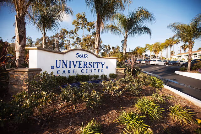 Entrance sign reading "University Care Center" with landscaping, palm trees, and a driveway with parked cars.