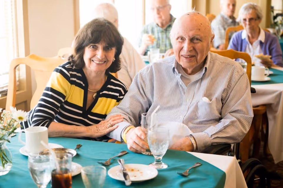 An elderly man and a woman sitting together at a dining table in a senior living facility, smiling at the camera. The table is set with glasses, plates, and utensils, and other elderly people are visible in the background also seated at tables.