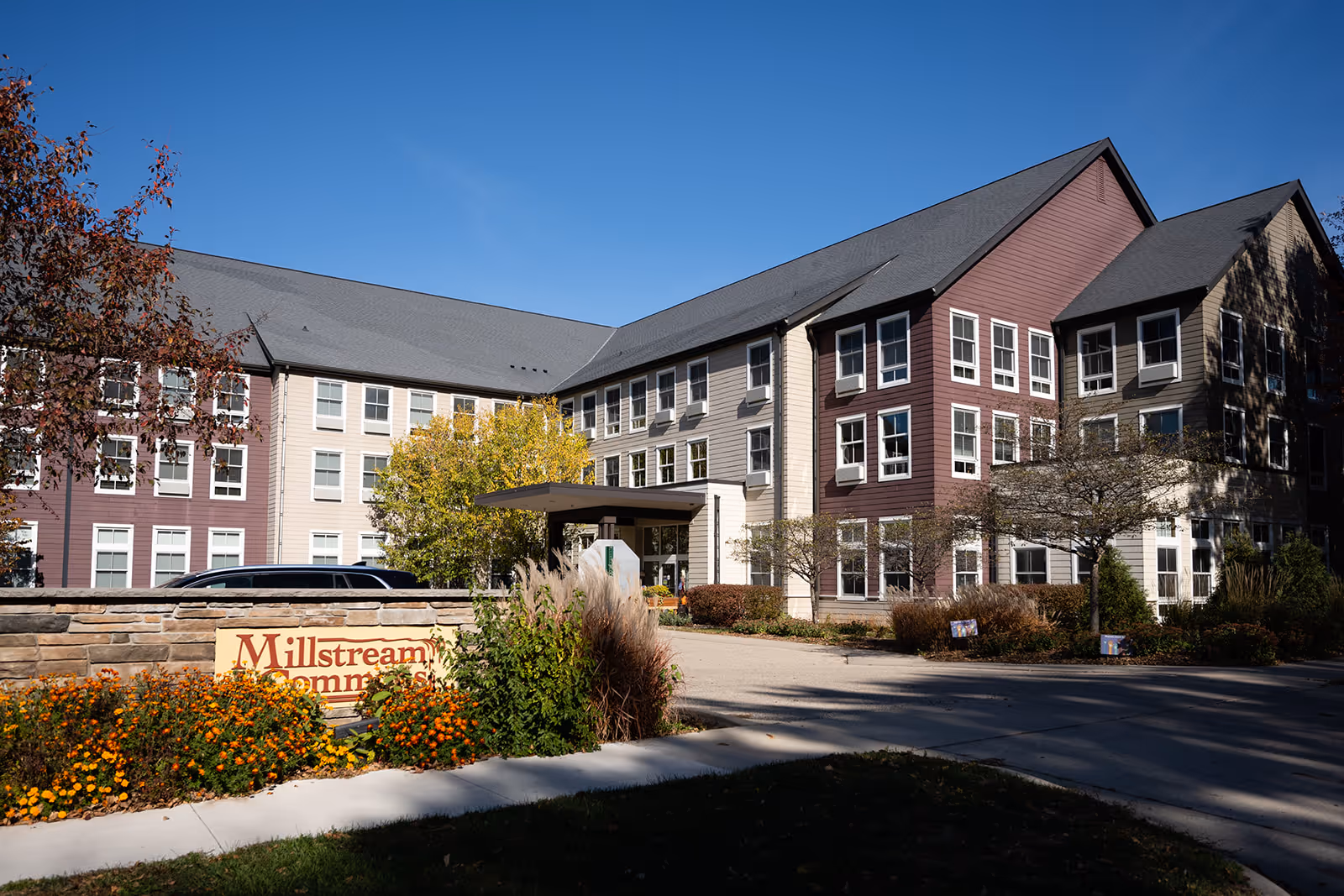 Front exterior of the Millstream Commons senior living building with an entrance canopy, landscaped flowerbeds, and a stone sign.