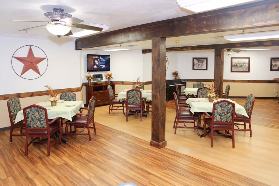 A dining room in a nursing and rehabilitation center with several tables covered with light green tablecloths and floral centerpieces. Each table is surrounded by chairs with floral upholstery and wooden frames. The room features wood flooring, wooden beams, a ceiling fan with a light, a wall-mounted TV, and framed pictures on the walls. A large decorative star is mounted on one wall.