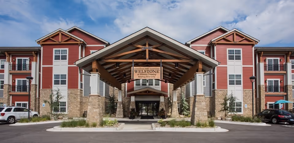 Front exterior view of The Welstone at Mission Crossing senior living facility with a covered entrance supported by stone pillars, red and beige building facade, and a clear blue sky.
