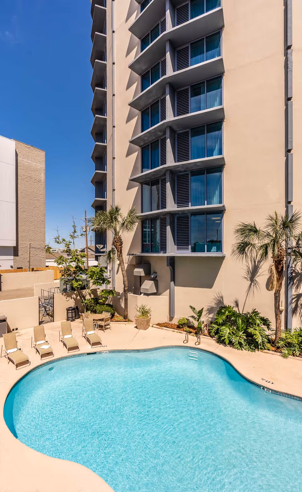 Outdoor swimming pool area with lounge chairs and palm trees next to a tall building under a clear blue sky.