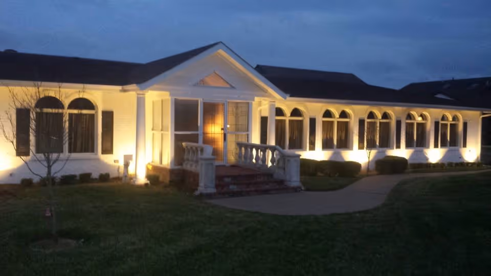 Exterior view of Mari De Villa Nursing Home at dusk, showing a single-story building with white walls, multiple arched windows, and warm outdoor lighting illuminating the facade and entrance with steps and white railings.