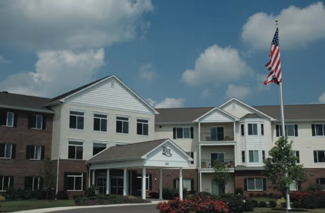 Exterior view of a multi-story senior living facility building with a covered entrance, landscaped bushes, and an American flag on a flagpole in front under a partly cloudy sky.