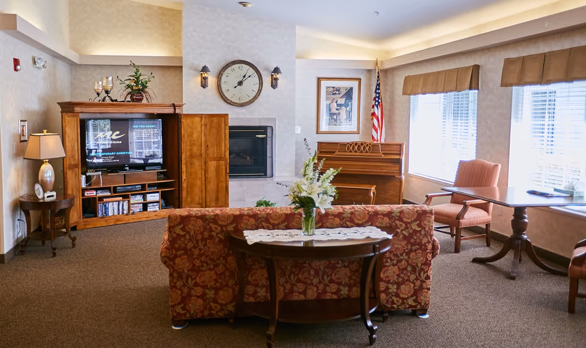 A cozy senior living common area with a floral patterned sofa facing a wooden entertainment center with a TV. There is a fireplace with a clock above it, a piano with an American flag beside it, and two armchairs near large windows letting in natural light. A small table with a vase of flowers is placed behind the sofa.
