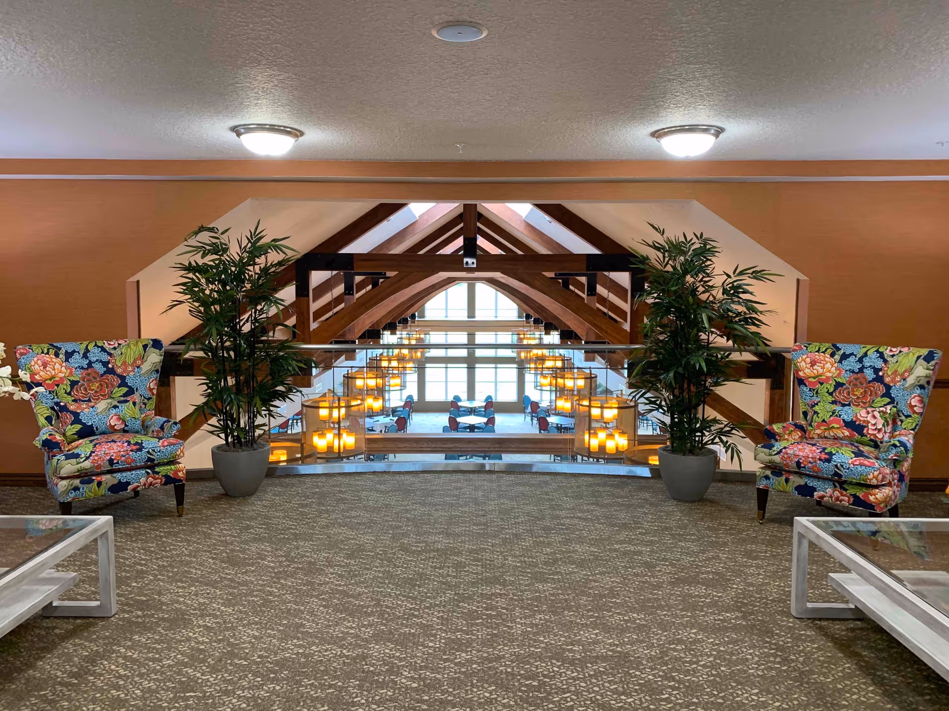 Interior view of a senior living facility lounge area with two colorful floral armchairs on either side, two potted plants, and a glass railing overlooking a dining area below with wooden beams and hanging lights.