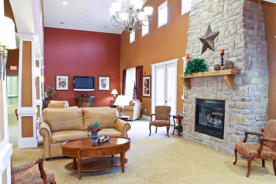 A spacious living room in an assisted living facility with beige sofas, patterned armchairs, a wooden coffee table with a flower vase and decorative items, a large stone fireplace with a star decoration and candles on the mantel, a flat-screen TV mounted on a red accent wall, and multiple windows letting in natural light.