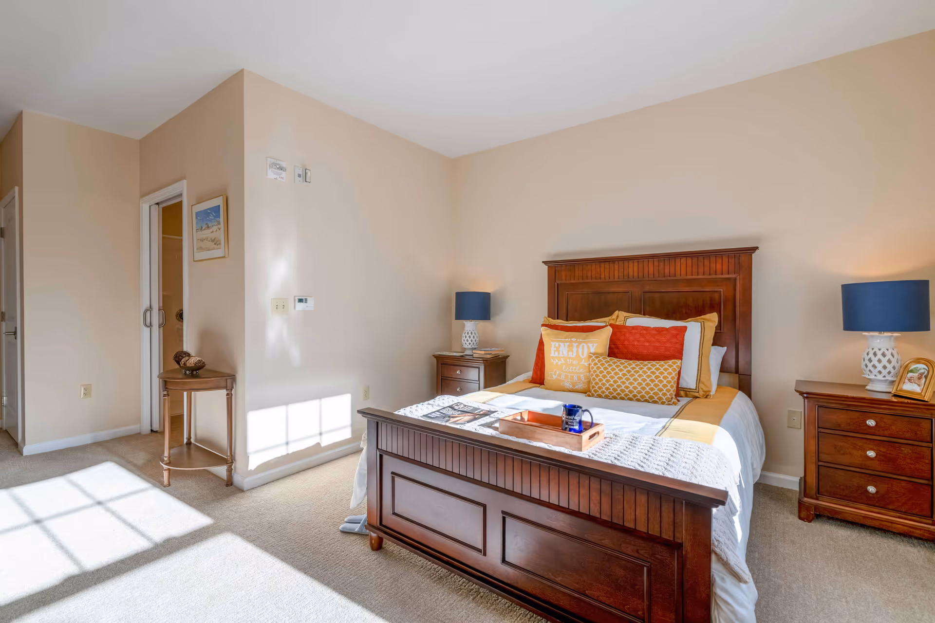 Sunlit bedroom with a wooden bed, bedside tables and lamps, decorative pillows, and a small side table by a doorway.