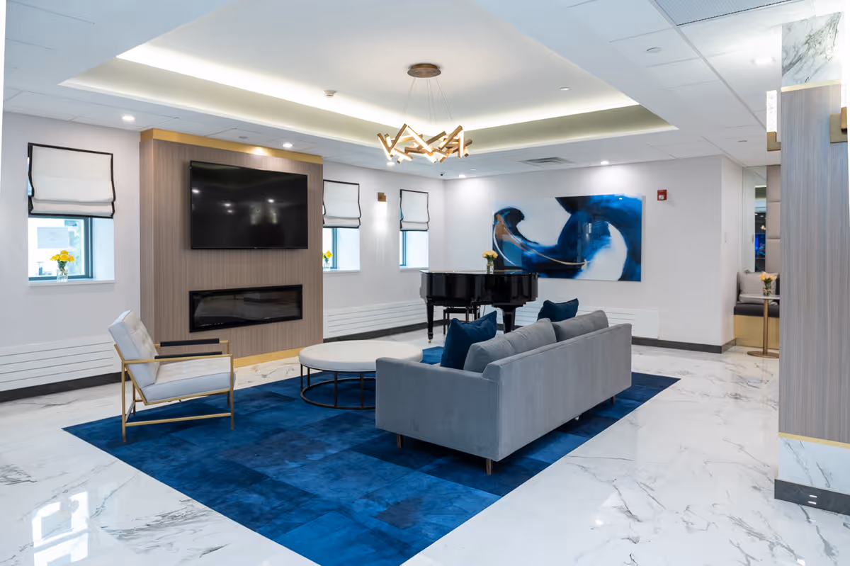 A modern living room area with a gray sofa and blue pillows on a large blue rug, a round white coffee table, and a single armchair with gold frame and white cushions. The room features a wall-mounted flat-screen TV above a built-in electric fireplace, three small windows with white roman shades, a black grand piano, and a large abstract blue and white painting on the wall. The floor is white marble with gray veining, and the ceiling has recessed lighting and a contemporary chandelier.