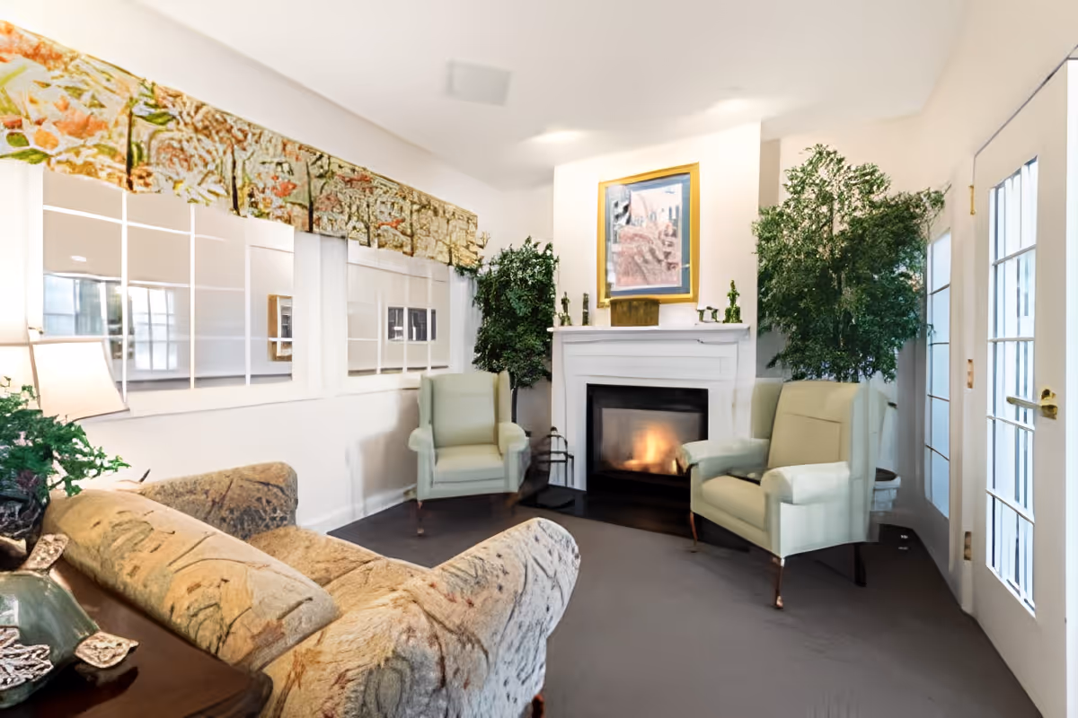 A cozy living room with a lit fireplace, two light green armchairs, a patterned sofa, large green plants, a framed painting above the fireplace, and a large mirror on the wall reflecting the room. There is a lamp on a side table next to the sofa and a glass door letting in natural light.