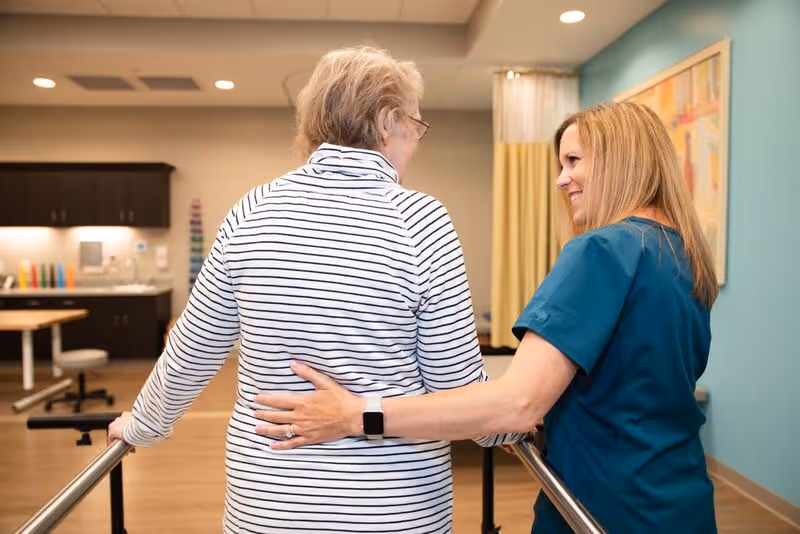 A healthcare worker in blue scrubs supports an elderly woman wearing a striped shirt as she walks between parallel bars in a rehabilitation or physical therapy room.