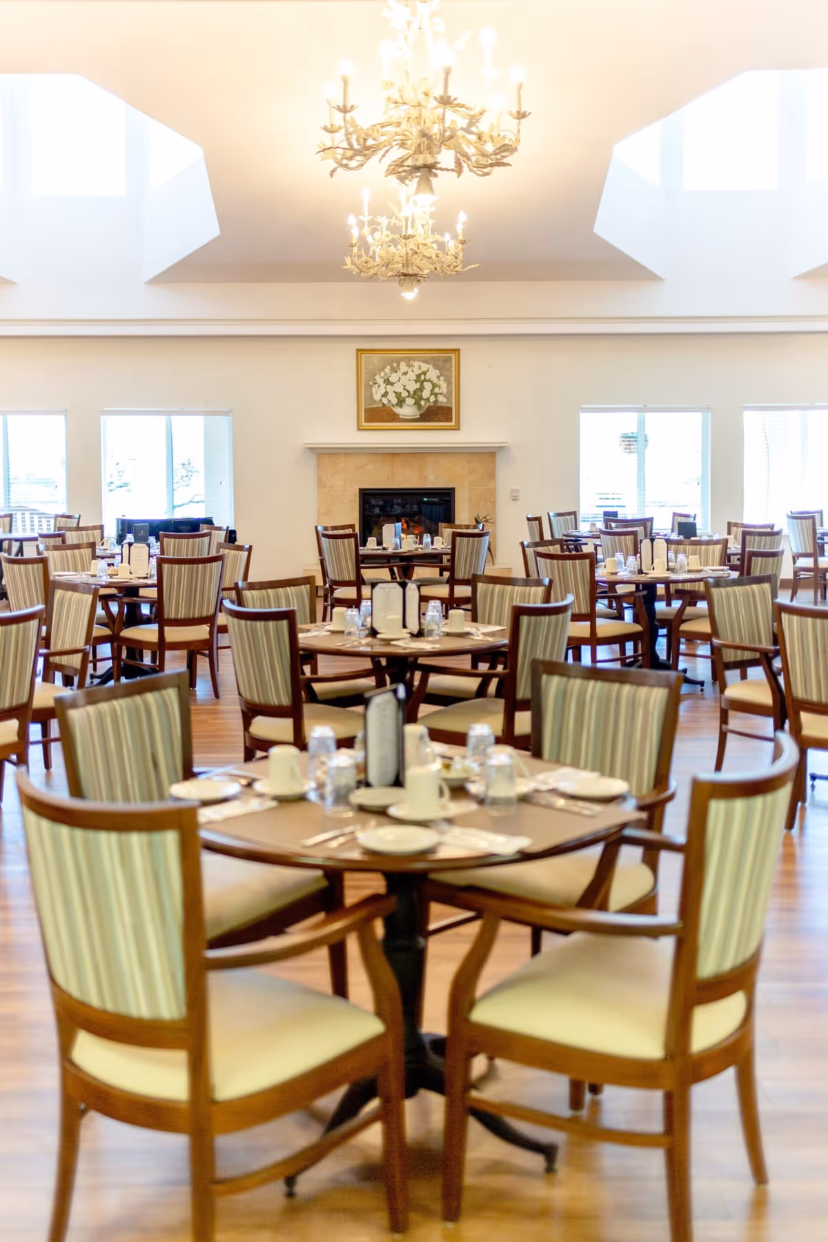 A bright and spacious dining room with multiple round tables set for meals. Each table has wooden chairs with striped upholstery, place settings including cups, plates, and utensils, and menus. The room features large windows allowing natural light, a fireplace with a floral painting above it, and two ornate chandeliers hanging from the ceiling.